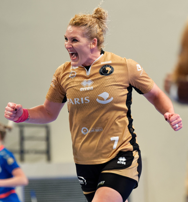 Eine Frau in brauner Bluse und schwarzen Shorts rennt und feiert auf einem Futsalplatz, mit zwei anderen Frauen im Hintergrund.
